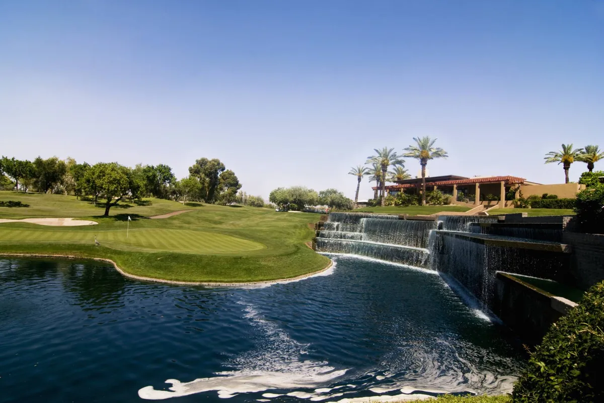 This image features a serene golf course with a glistening waterfall, palm trees, and a residential building subtly visible in the background. The scene is bathed in warm light, highlighting the peacefulness of the natural setting and the proximity of recreational amenities.