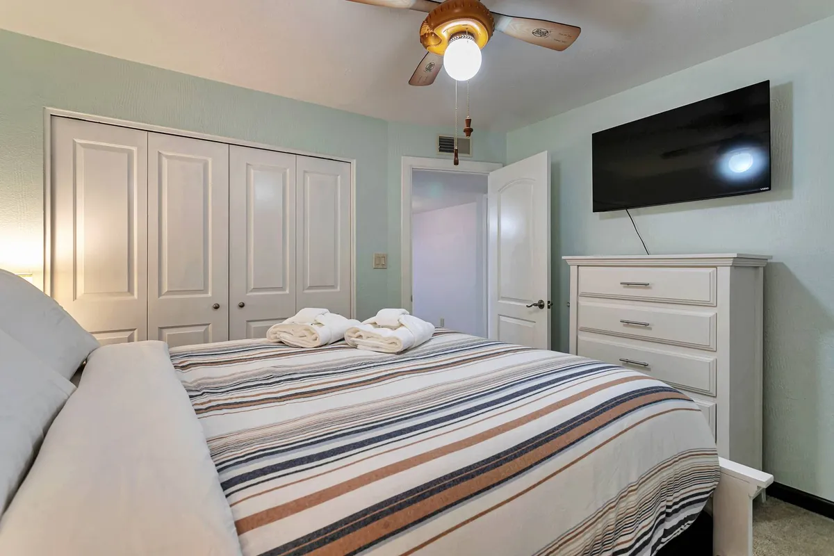 A spacious master bedroom featuring a king bed with striped linens, a wall-mounted TV, a dresser, and a ceiling fan. Natural light fills the space, emphasizing comfort and tranquility.