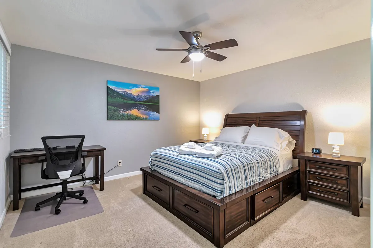 A serene guest bedroom with a bed dressed in striped bedding, a desk and chair perfectly suited for a dedicated workspace, a nightstand with a lamp, and tasteful wall art. Morning light adds to the calming ambiance.