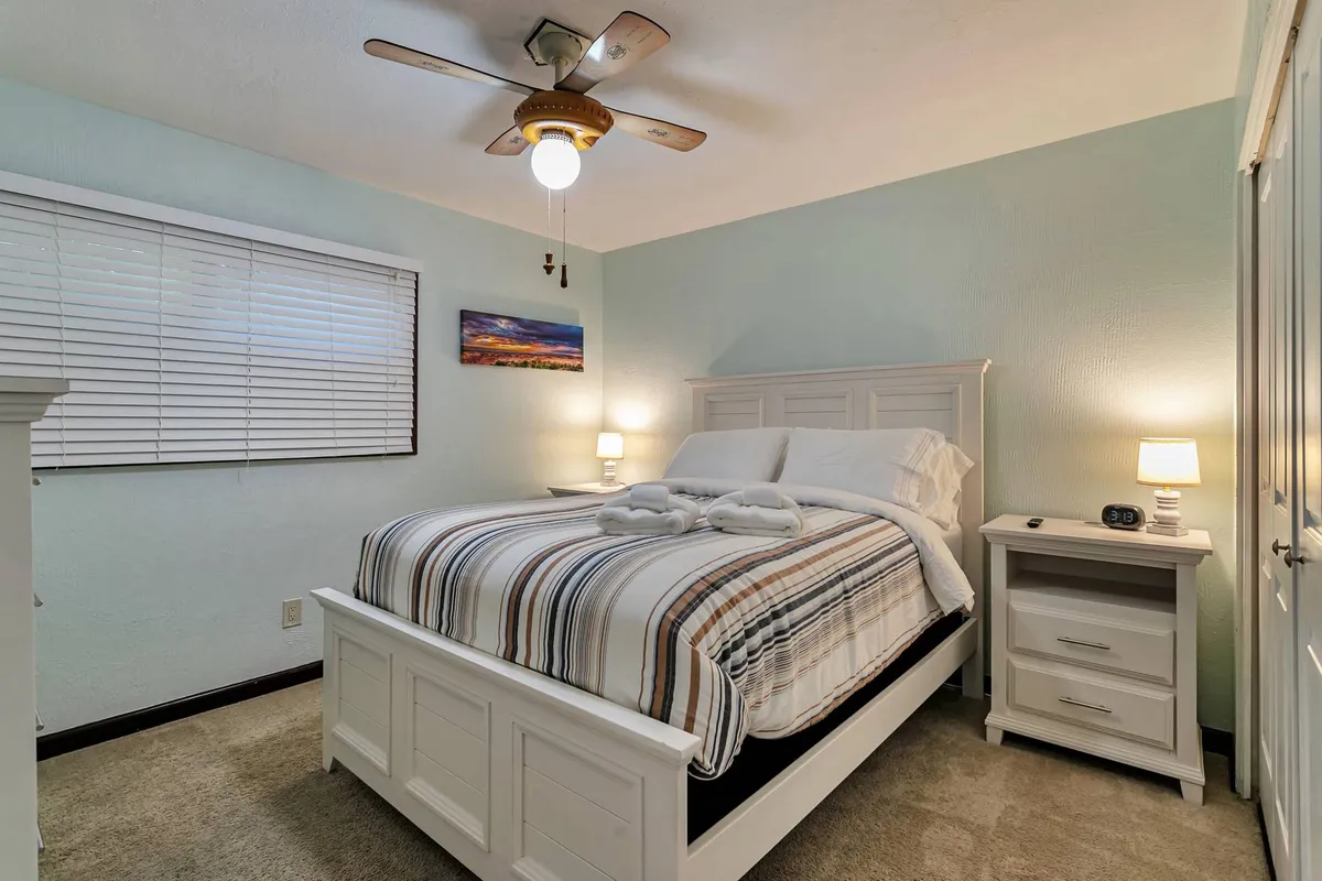 A tranquil guest bedroom featuring a queen bed with striped bedding, matching nightstands with lamps, a ceiling fan, and decorative wall art. Soft natural light creates an inviting atmosphere.