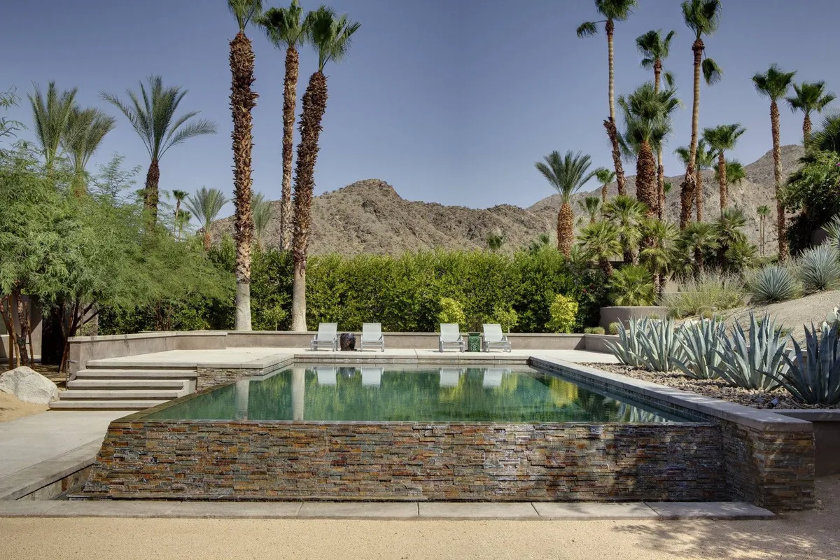 A serene view of the swimming pool framed by palm trees and a dramatic mountain backdrop, with plenty of lounge chairs inviting guests to relax. The sunset sky adds a resort-like tranquility to the scene.