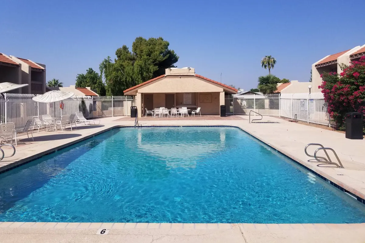A bright and relaxed view of the community swimming pool with ample poolside seating, a clubhouse in the background, and sunny skies overhead. The area is set up for swimming, lounging, socializing, and enjoying the Arizona weather.