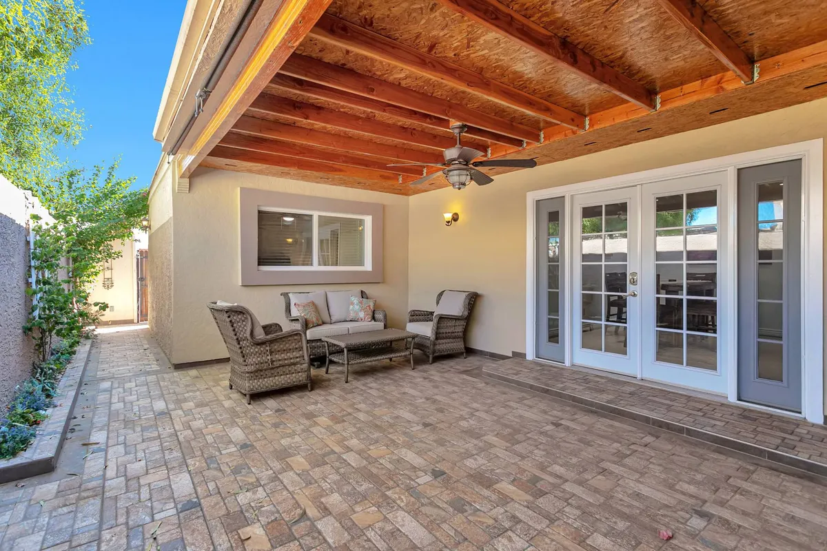 An inviting exterior patio area featuring a cozy wicker seating arrangement, sliding glass doors connecting to the home, overhead wooden ceiling, and lush greenery lining a stone pathway. The space feels welcoming, ideal for enjoying Arizona's sunshine or sharing outdoor meals with family and friends.