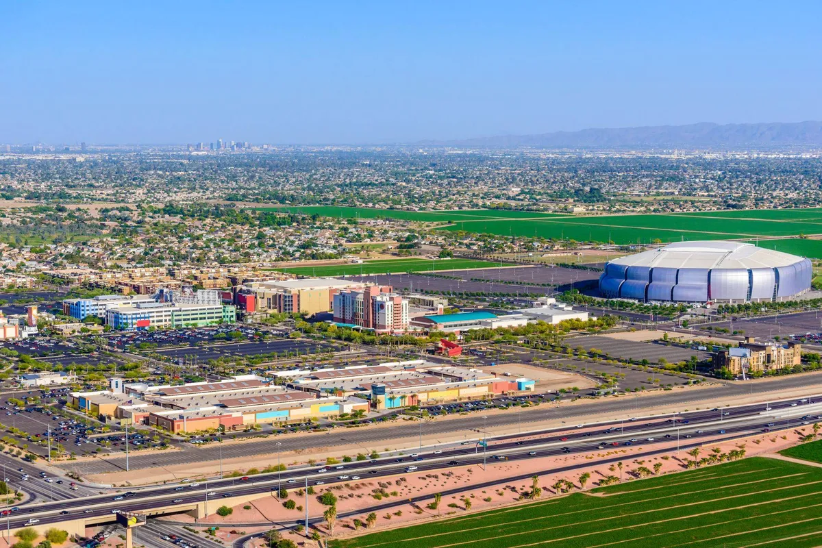 A wide exterior view showing a bustling shopping center, open green fields, a major stadium, and a nearby highway, capturing the dynamic energy of the neighborhood and its proximity to local attractions and events.