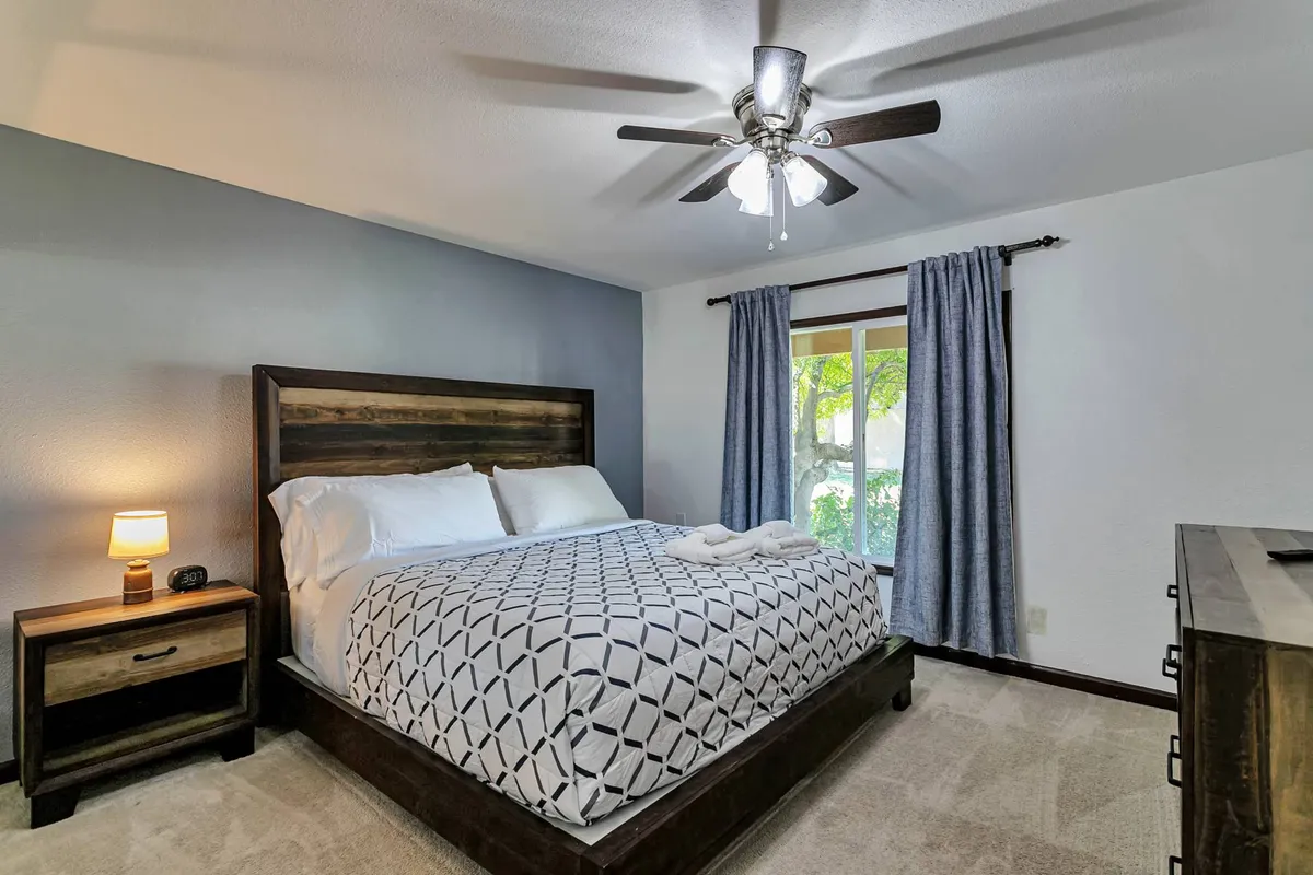 A serene bedroom featuring a comfortable bed with soft linens, a nightstand beside the window, elegant curtains, and a ceiling fan overhead. Sunlight filters through the window, creating a tranquil, inviting atmosphere perfect for unwinding after a day of activities.